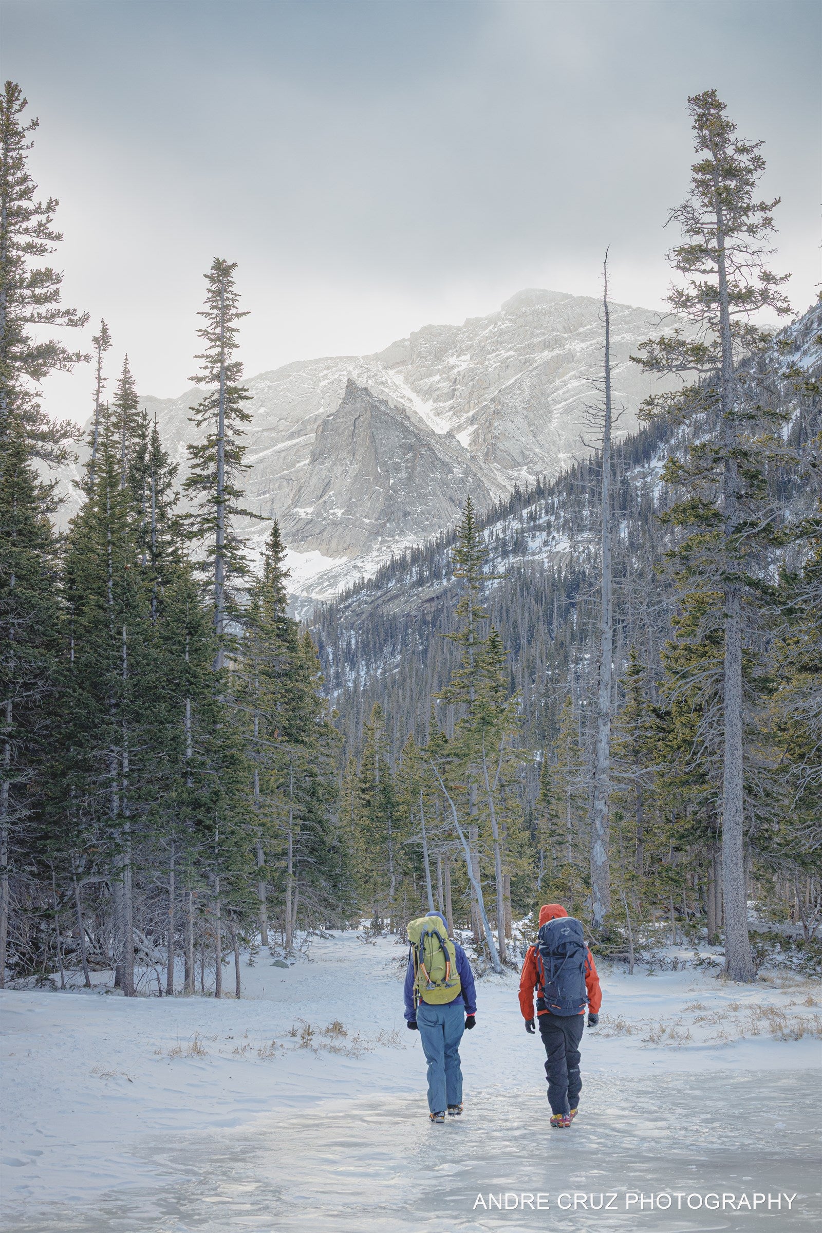 Two hikers walking away across a frozen alpine lake toward snow-covered peaks, Rocky Mountain National Park