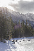 Sun breaking over a spired ridge with backlit pines above a frozen lake in winter
