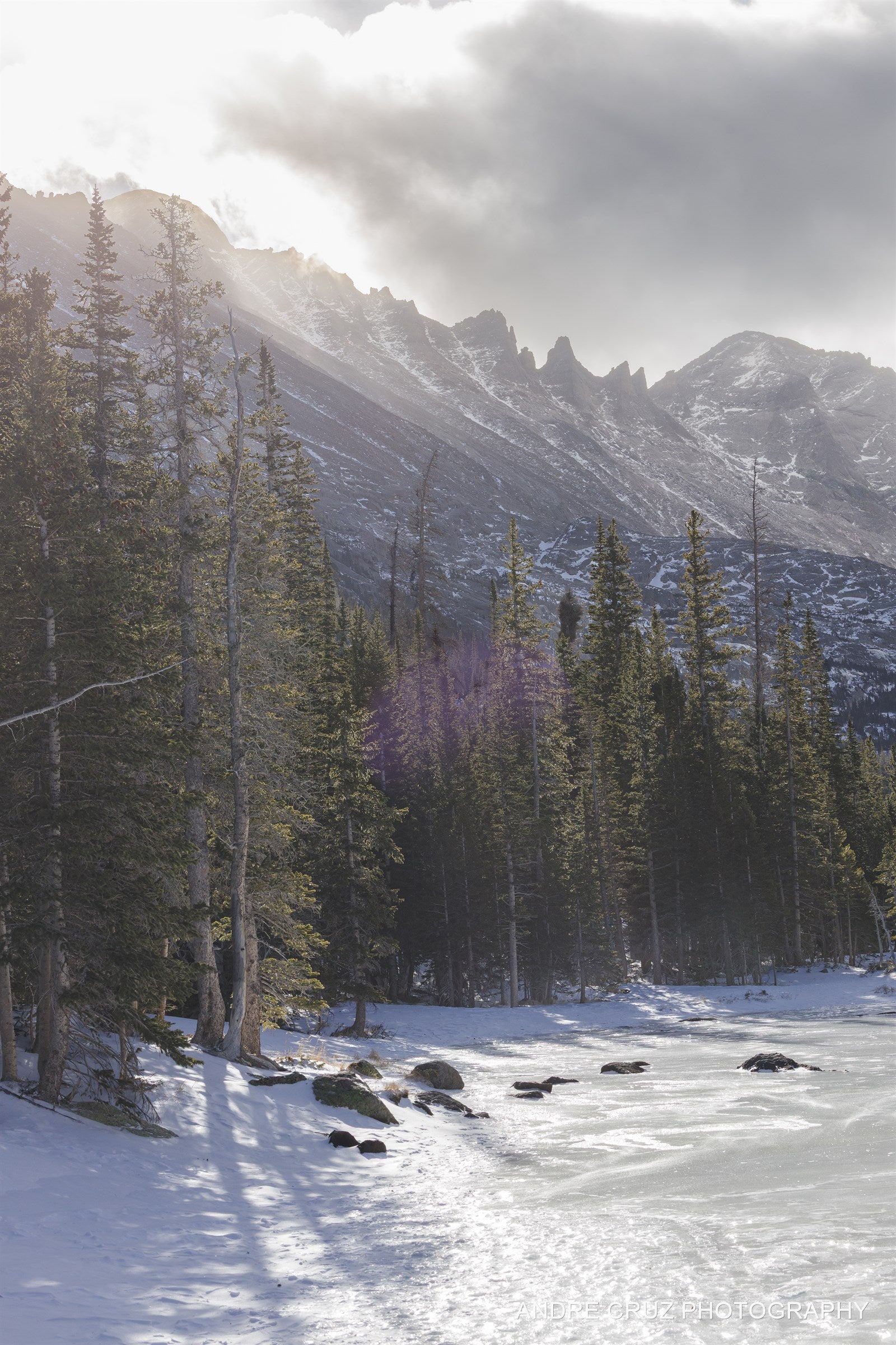 Sun breaking over a spired ridge with backlit pines above a frozen lake in winter