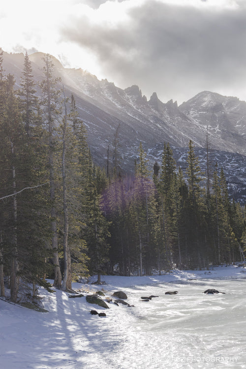 Sun breaking over a spired ridge with backlit pines above a frozen lake in winter