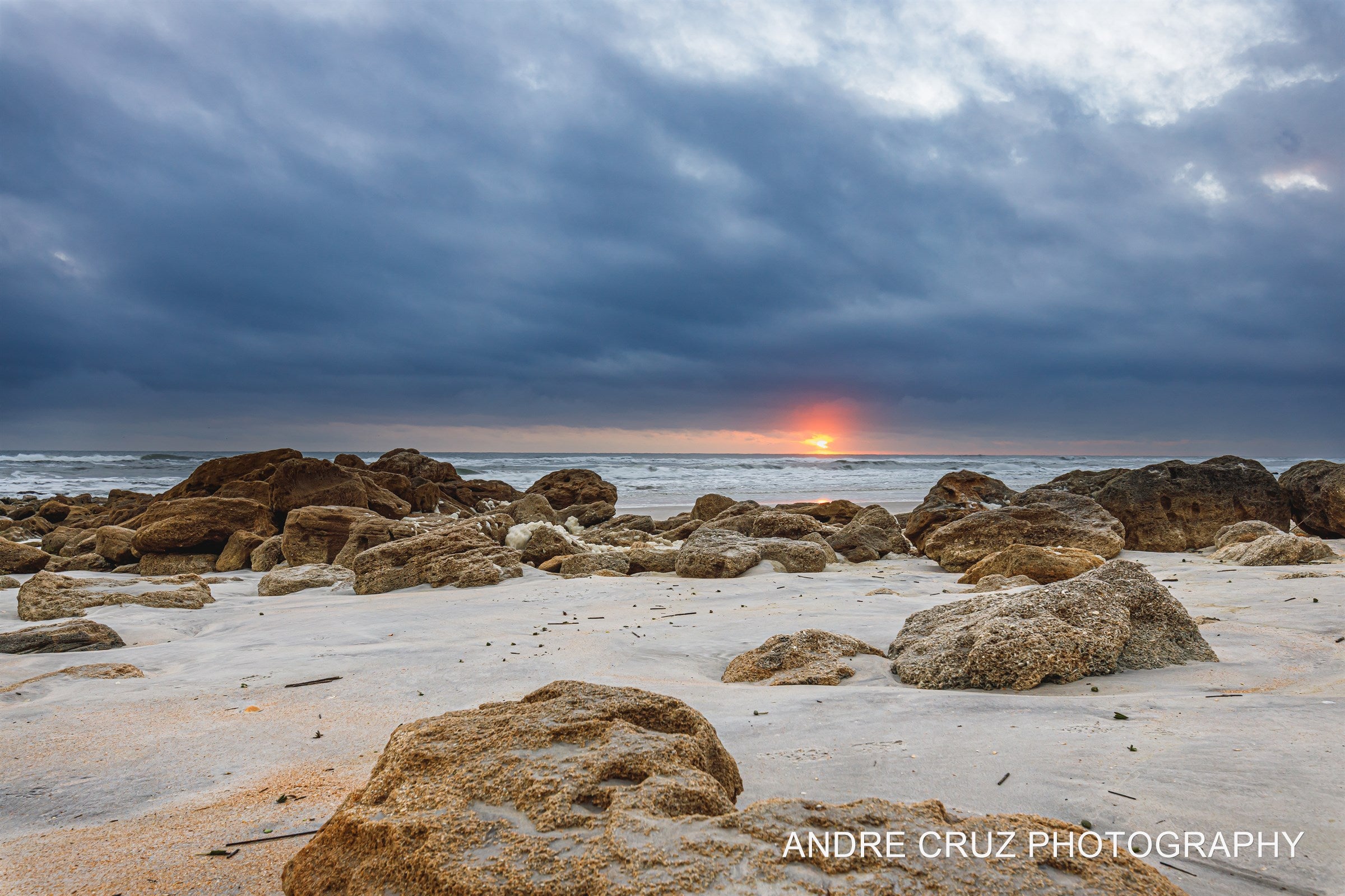 A low sun breaking through a heavy grey cloud bank over coquina rock and wet sand, Atlantic coast of Florida