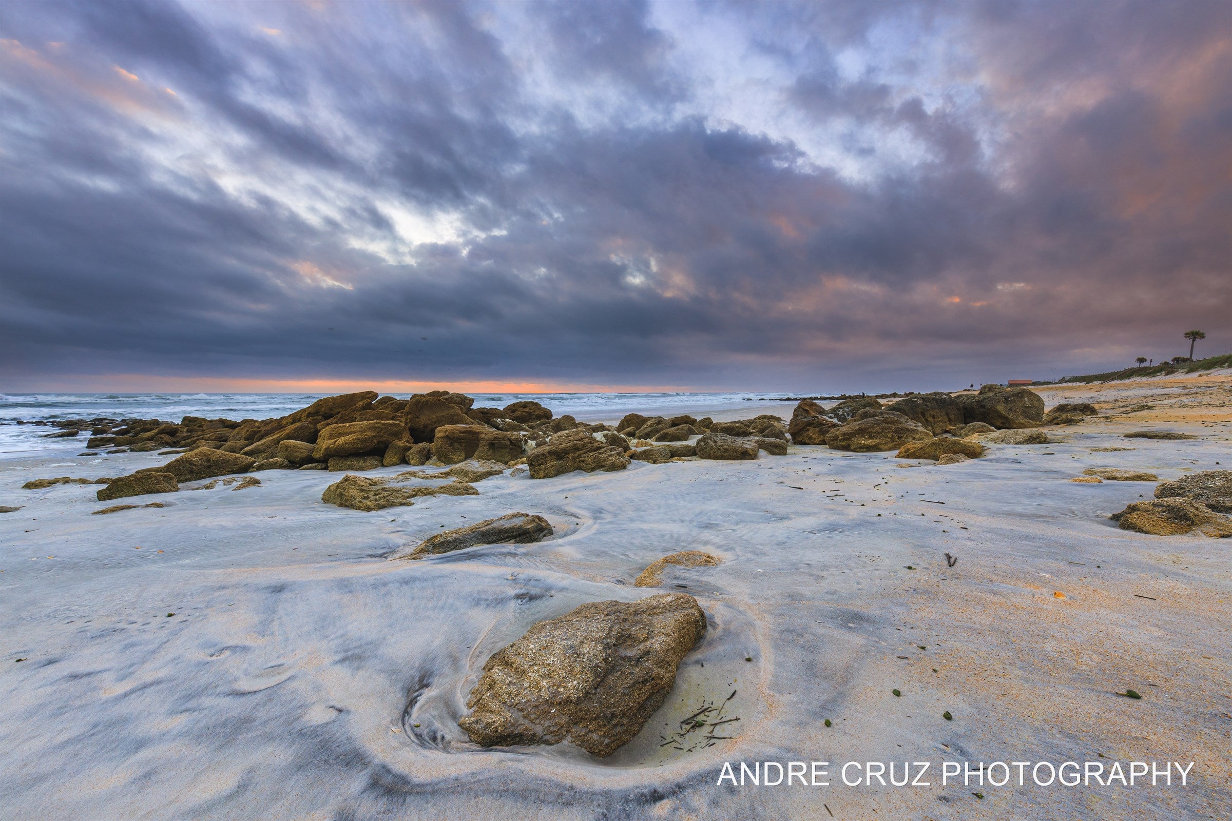 Long-exposure water swirling around a rock on the coquina flats under a pink and violet dawn sky, Florida