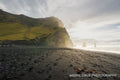 Low sun across the black sand at Reynisfjara, a green headland on the left and the Reynisdrangar sea stacks offshore