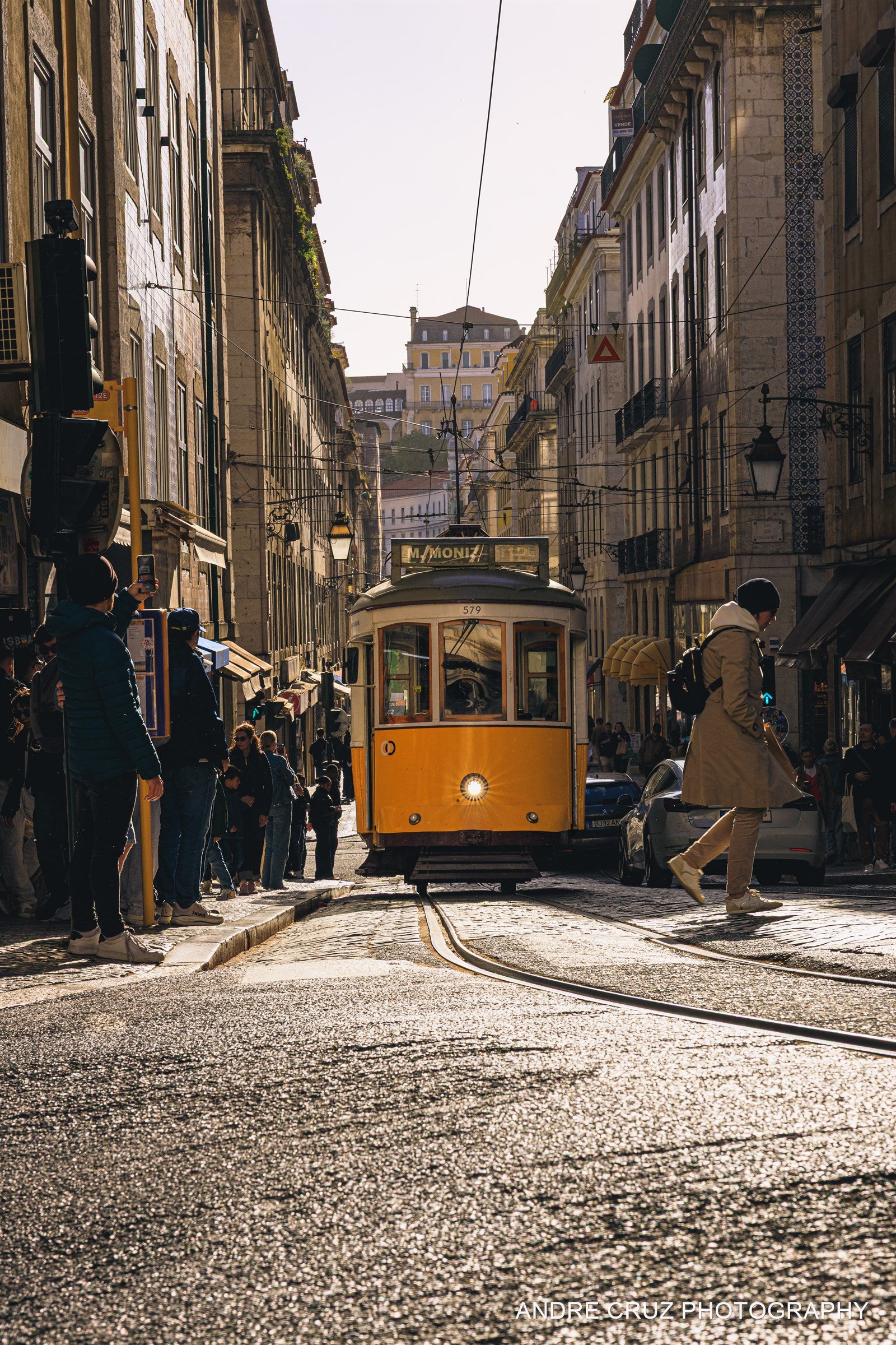 A yellow tram climbing a steep cobbled hill in Lisbon with pedestrians alongside, Portugal