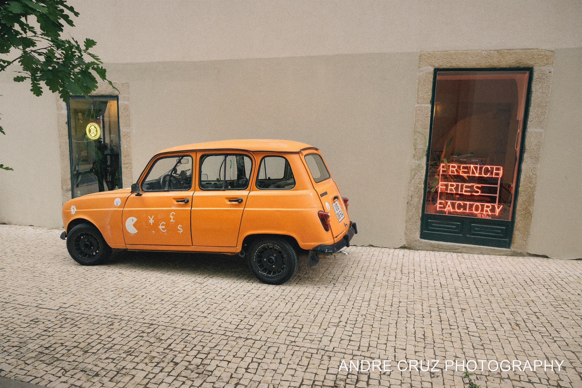 An orange Renault 4 parked on cobbles against a white wall beside a red French Fries Factory neon sign