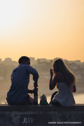 Two people sitting on a wall at a viewpoint above a hazy city at golden hour, seen from behind
