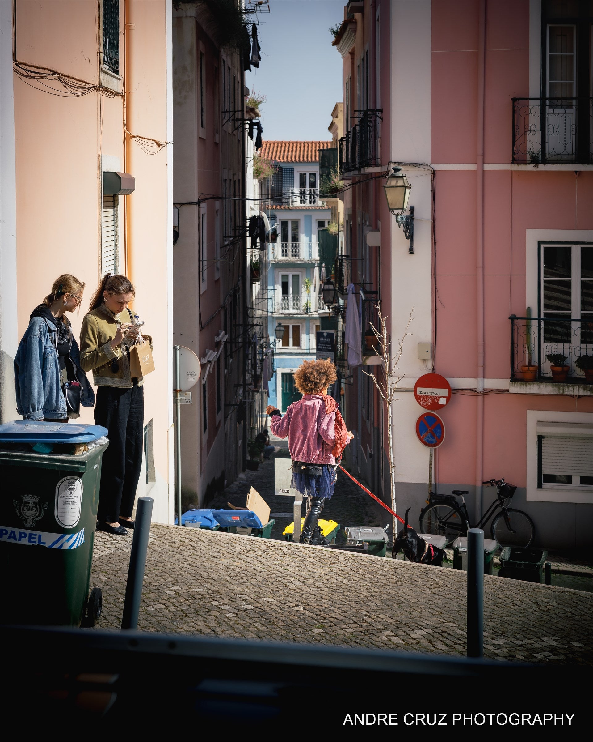 Two women talking on a steep pink-walled Lisbon street while a child pushes a scooter downhill